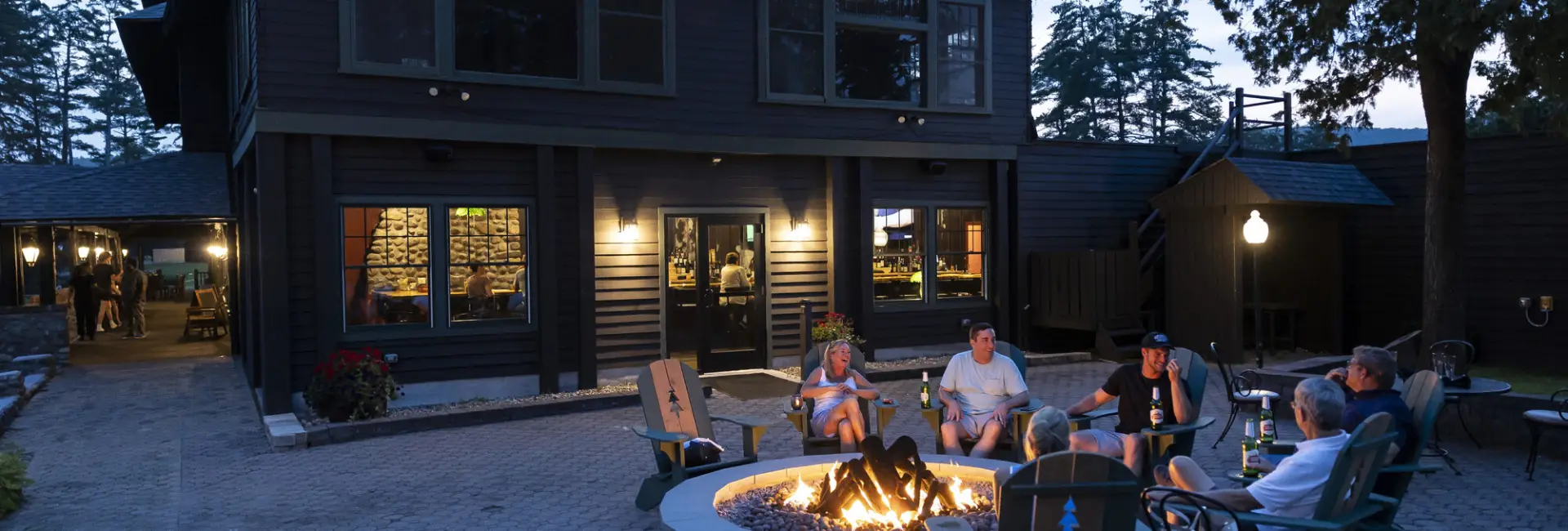 A group of people sit at an outdoor stone firepit at a lodge in summer. 