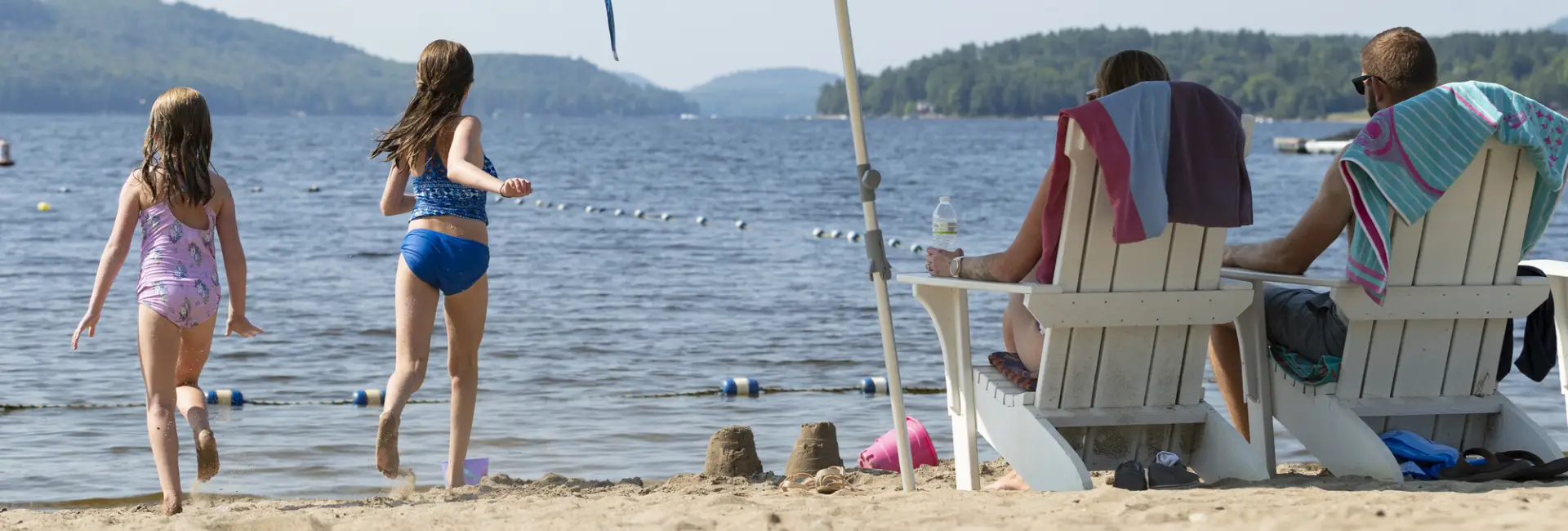 Two children laugh and play on a beach while their parents sit in beach chairs. 