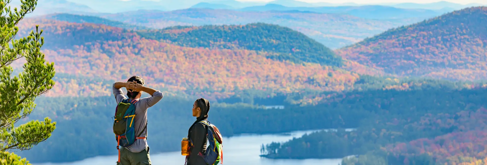 A man and woman stand on top of a mountain in fall overlooking a lake and mountains.