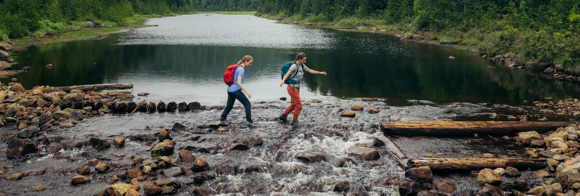 Two women walk across a rushing stream in the middle of the forest. 