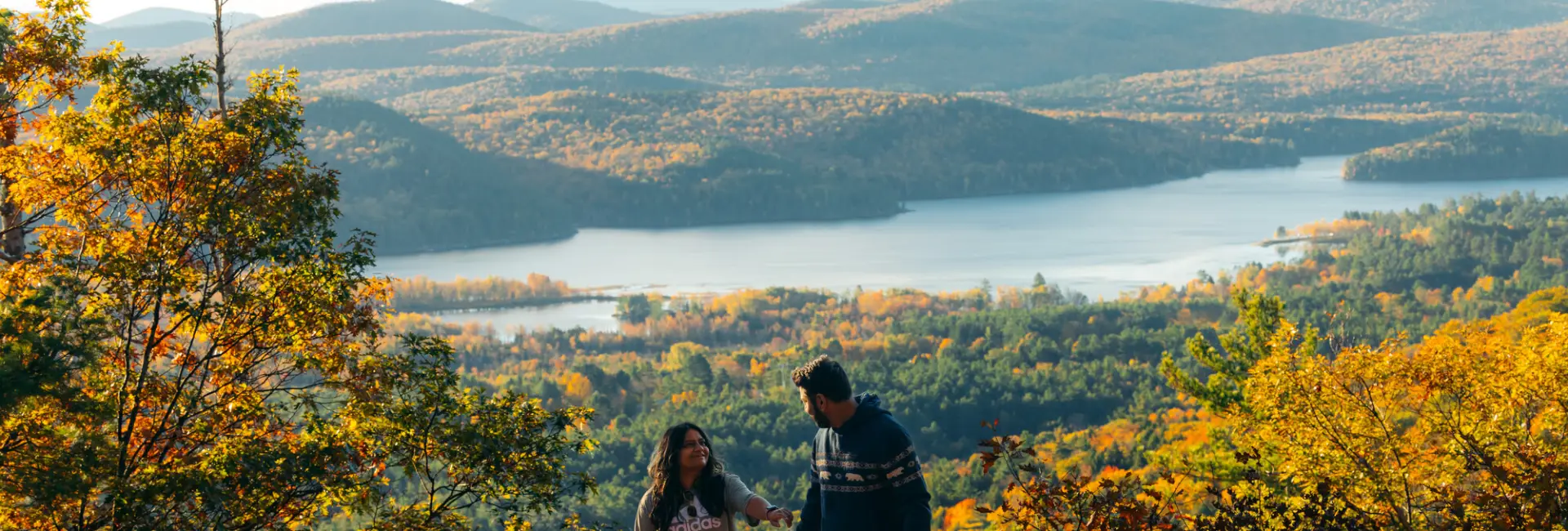 A man and woman hike up a fall mountain with a lake down below. 