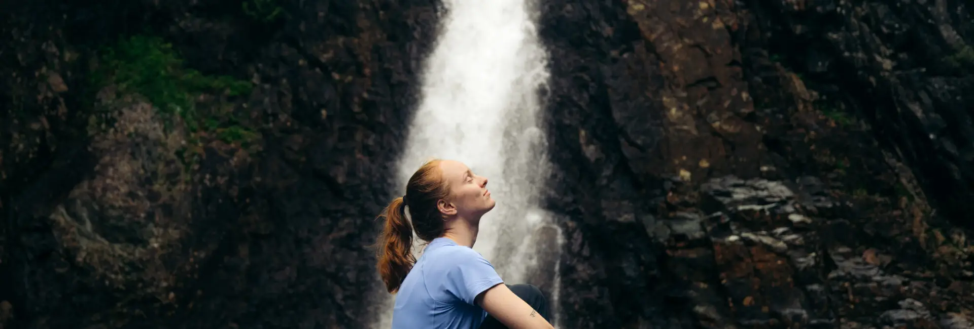 A woman sits at the base of a waterfall.