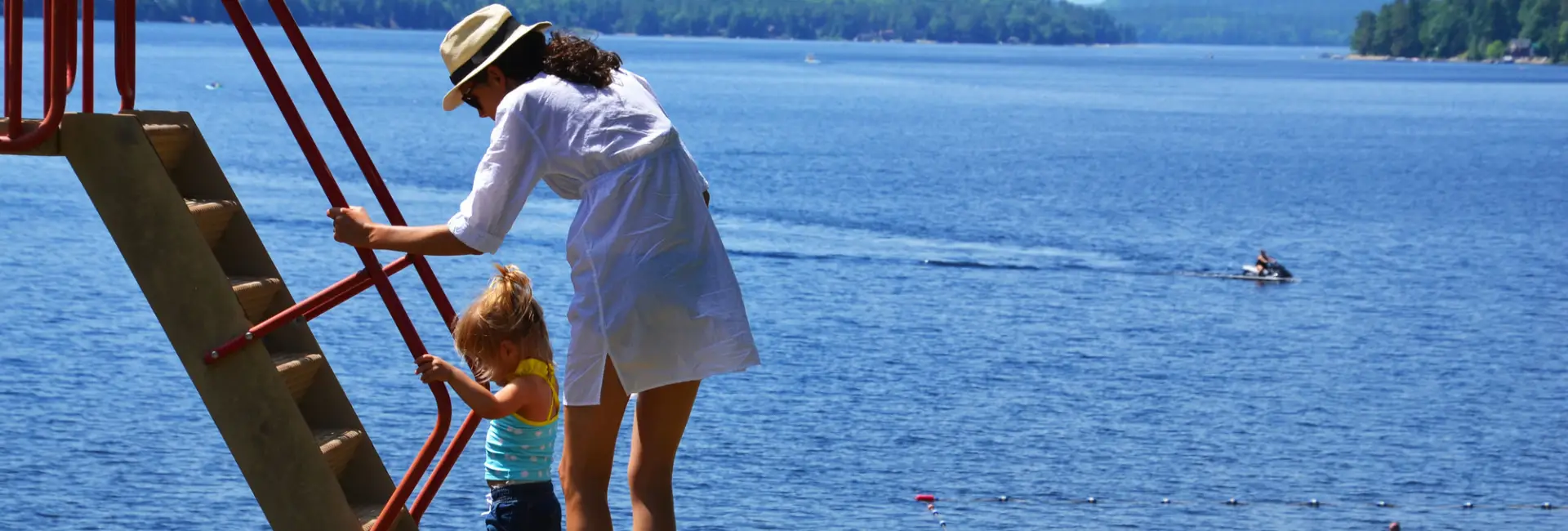 A woman in a dress and fedora helps her toddler climb stairs on a playground at a beach. 