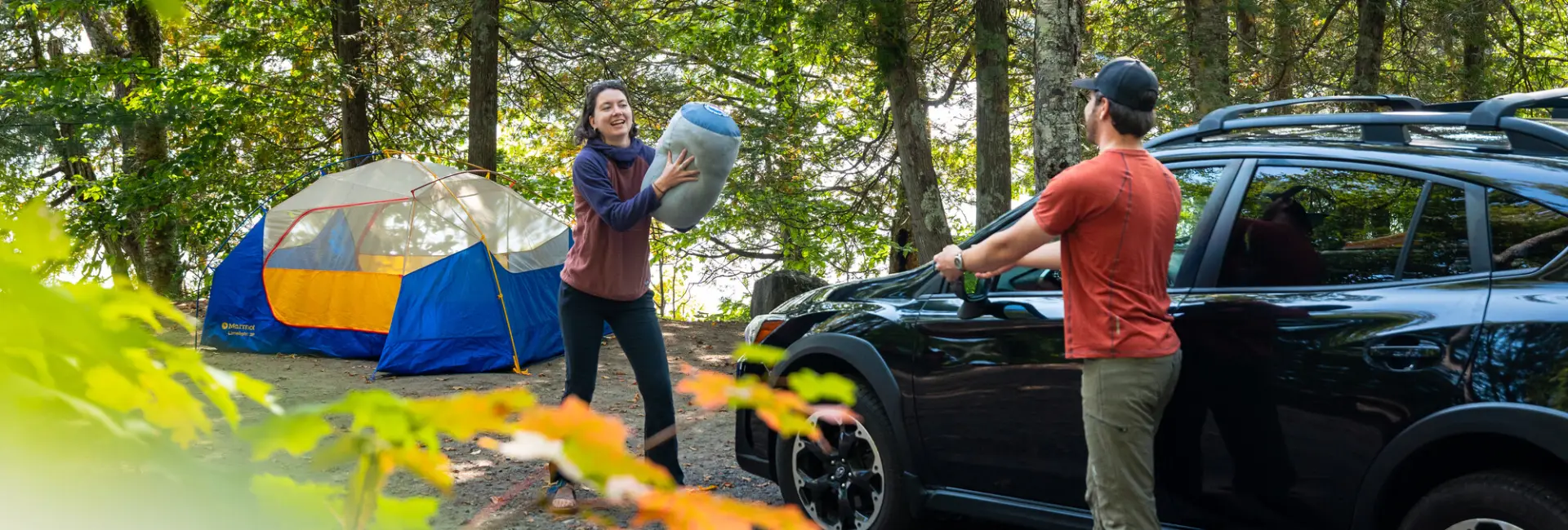 A woman throws a sleeping bag to a man next to a car and tent.