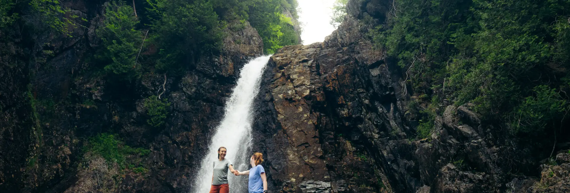 two women stand beneath a waterfall in summer. 