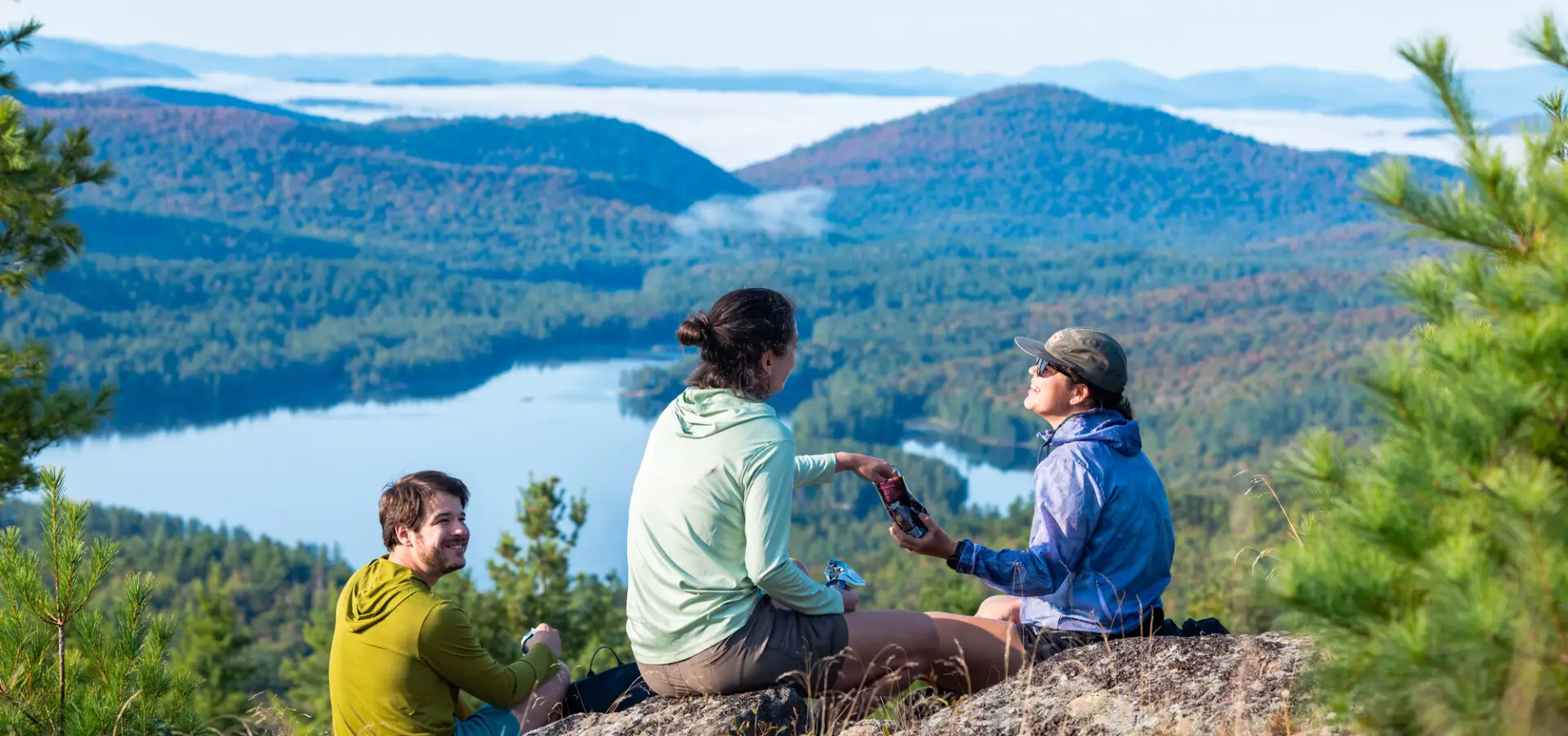Three adults sit on a rocky summit overlooking a lake and forested mountains on a summer day.
