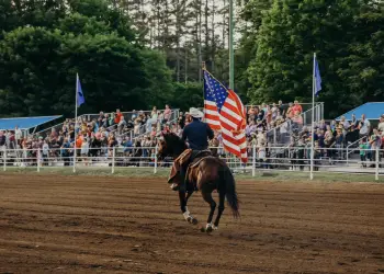 A man carrying an American Flag rides a horse down the fairway while spectators look on.