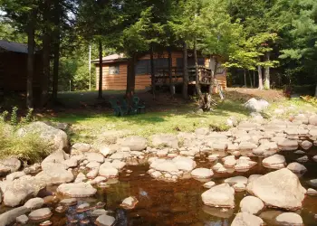 An image of a quaint cottage surrounded with trees and a rocky rivers edge.