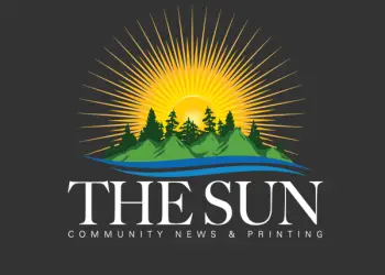 The Sun Logo of a large yellow sun,  a body of water,  a small green island with tall dark pine trees
