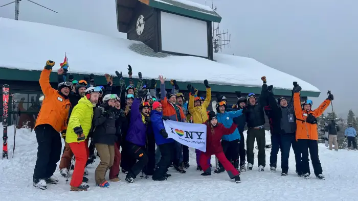 A large group of skiers and snowboarders in winter gear cheer and wave while holding an I Love NY LGBTQ+ pride flag.