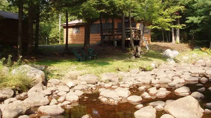An image of a quaint cottage surrounded with trees and a rocky rivers edge.