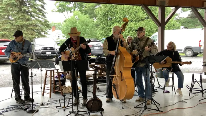 A bluegrass band performs under a covered pavilion. Five musicians play a variety of string instruments including a mandolin