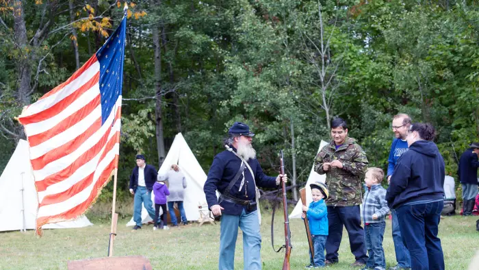 A man in a Union soldier uniform carrying a musket and talking with people attending the event. An American flag of that era is flying on a pole and there are white canvas tents in the background