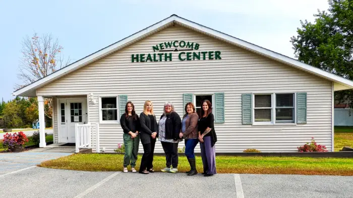 Five smiling women stand out side of the Newcomb Health Center building