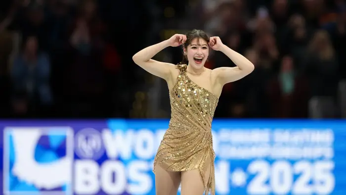 Male figure skater in brown long sleeve and long pant spandex suit performs a crouched move on ice