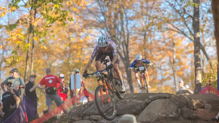 Cross-country mountain bike races rides over large rock while people look on during UCI MTB World Cup race in October