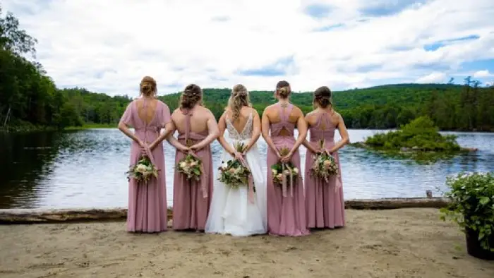 Bridal party stands in the sand by the water holding bouquets behind their backs.