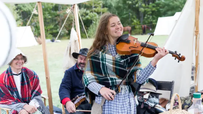 A young woman is playing a fiddle dressed in the Civil War era with others dressed in period clothing sitting in the background