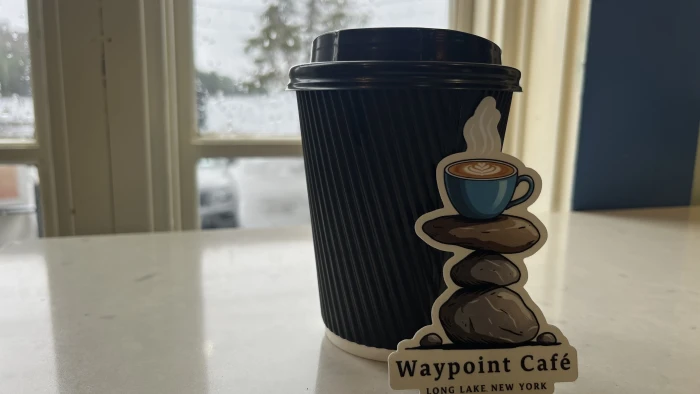 A hot cup of coffee on a table in a to-go cup with custom Waypoint Cafe sticker leaning against it.