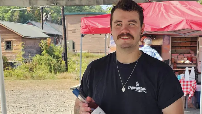A man smiles at the camera while holding a bottle of Springbrook Distillery's Adirondack Straight Bourbon.  He is standing behind a table displaying various bottles of moonshine and bourbon