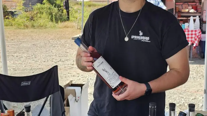 A man smiles at the camera while holding a bottle of Springbrook Distillery's Adirondack Straight Bourbon.  He is standing behind a table displaying various bottles of moonshine and bourbon