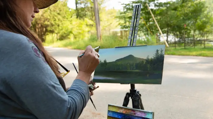 A person painting a lake scene outdoors on a small canvas