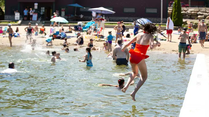 A girl in a red swimsuit jumps into a lake at a crowded beach.  Many people are swimming
