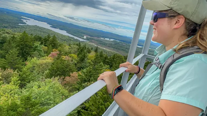 A hiker looks out of the fire tower on Buck Mountain over a forested landscape with a lake below.