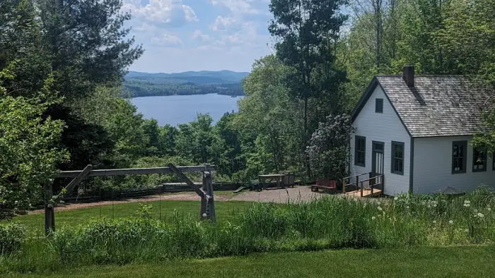 Blue Mountain Lake under billowing white clouds across a lush green lawn in front of a quaint white cabin.