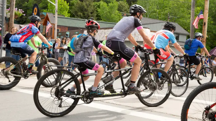 Cyclists race on a sunlit gravel road surrounded by lush green trees. The lead riders appear focused and determined