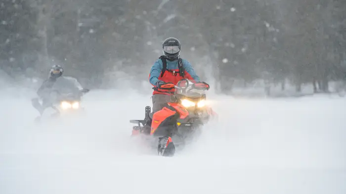 Two people ride snowmobiles across a snowy