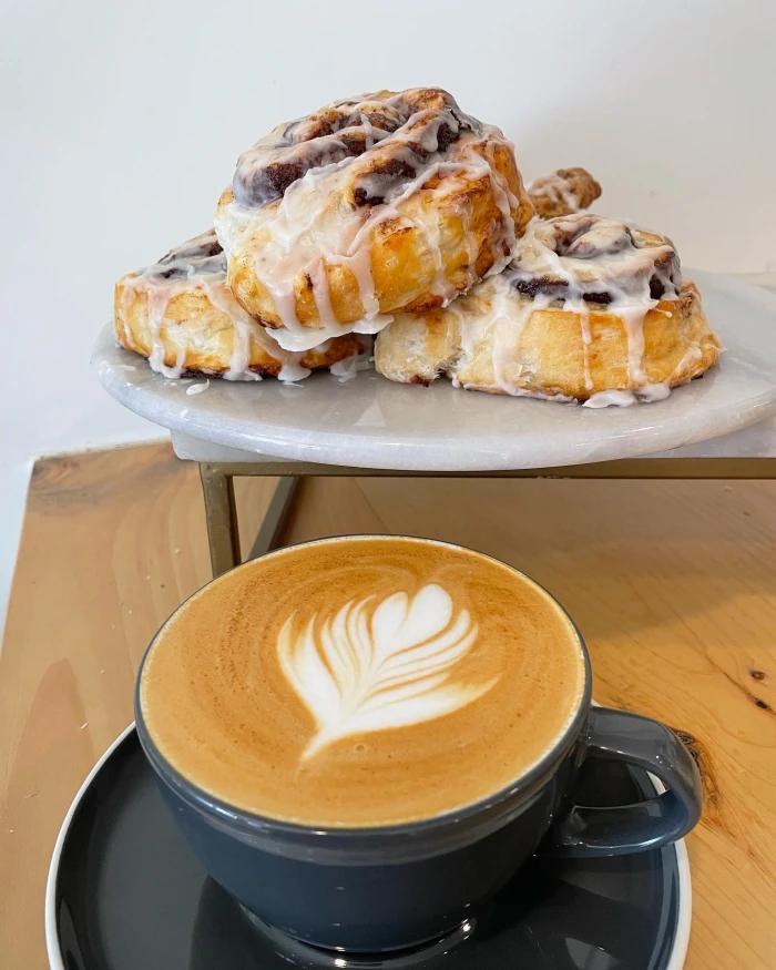 A foamy coffee drink in front of a platter of cinnamon rolls.