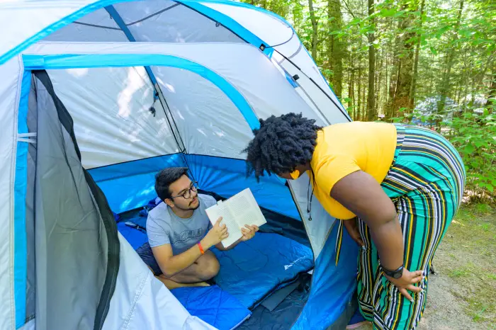 A woman bends over outside a tent as a man inside the tent shows her something in a book