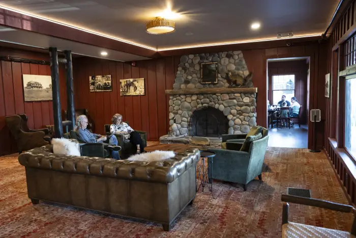 People read in an Adirondack-themed hotel lounge near a stone fireplace. A family dines in the restaurant in the background.