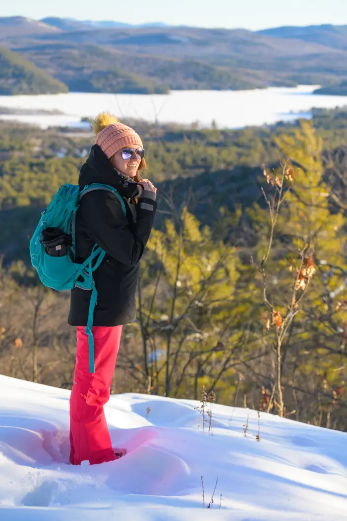 A woman in snow pants and jacket smiles at the top of a sunny snow covered mountain.
