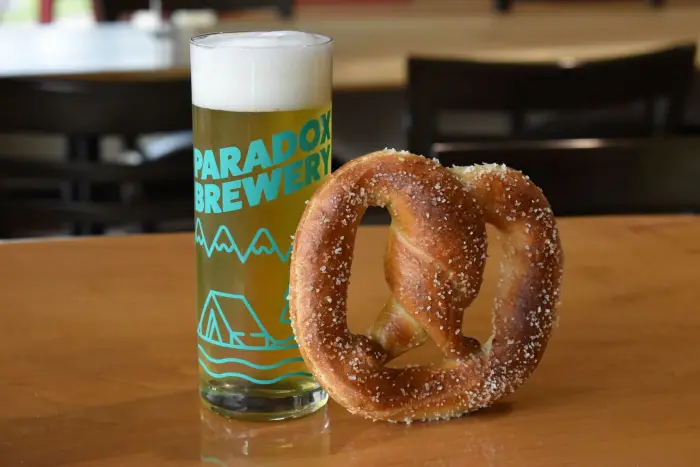 A tall&#44; narrow glass of beer sits on a wooden bar next to a big soft pretzel