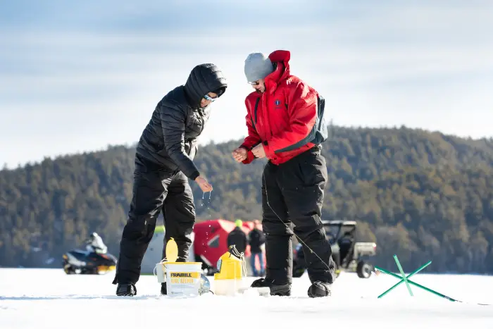 Two people look into a hole in the ice as they set up their ice fishing gear.