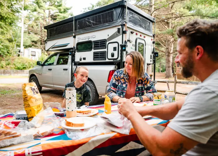 A man and woman have lunch with a young boy with a truck camper in the background.