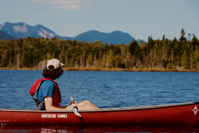 A paddler sitting in a red canoe looks across a body of water toward distant Adirondack peaks