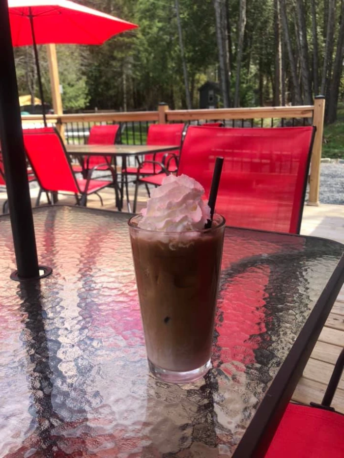 An iced coffee in a tall glass sits on a table in an outdoor dining area featuring bright red umbrellas and chairs.