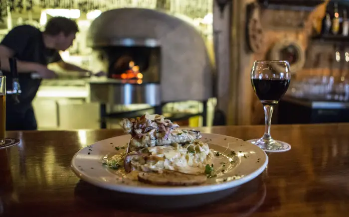 A plate of food and a glass of wine on a bar top with a man in the background putting food into a wood-fired oven