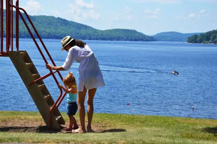 A woman in a swim coverup helps a small child on playground equipment&#44; with a large lake in the background.