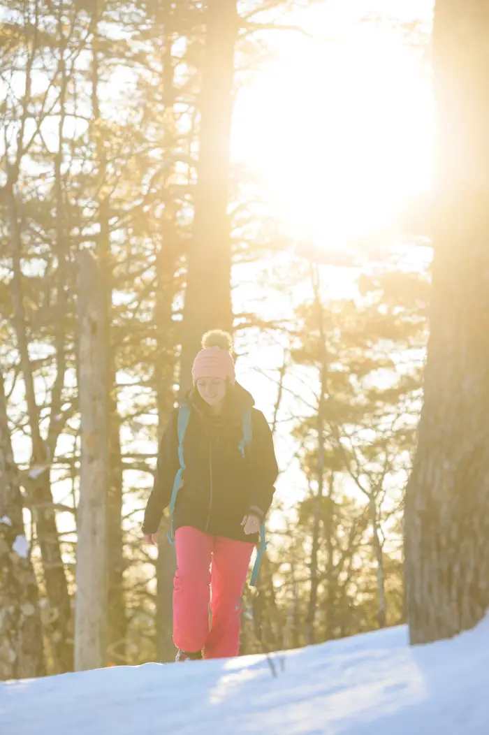 Woman walking on a snowy trail with sun shining through the trees.