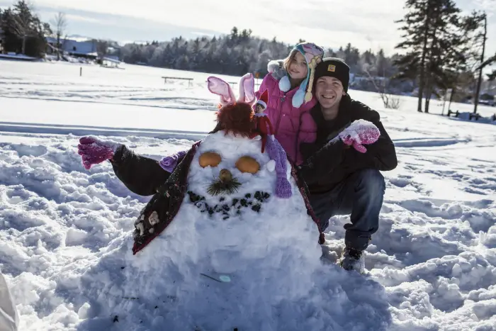 A young girl and her dad standing behind a snowman.