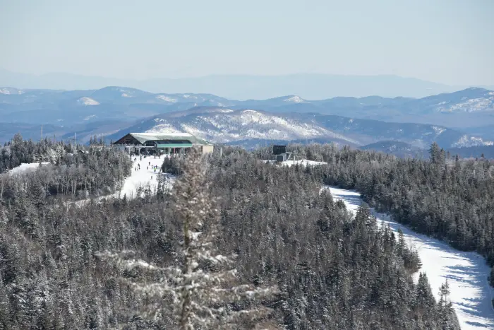 Snowy mountain landscape with a ski lodge and slopes surrounded by evergreen trees&#44; overlooking distant snow-covered peaks under a clear blue sky.