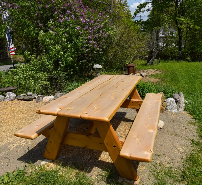 A light tan cedar picnic table near green grass with a purple lilac in bloom behind.