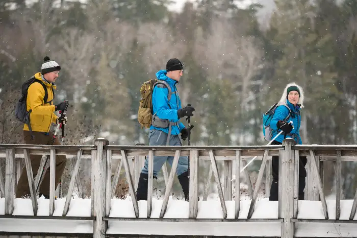 three people on snow shoes cross a snow covered bridge.