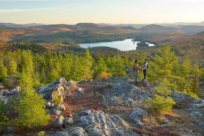 Two hikers stand on a mountain with a blaze of fall colors and a lake below.