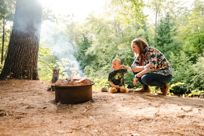 A woman and a child crouch near a metal fire pit at a wooded campsite.