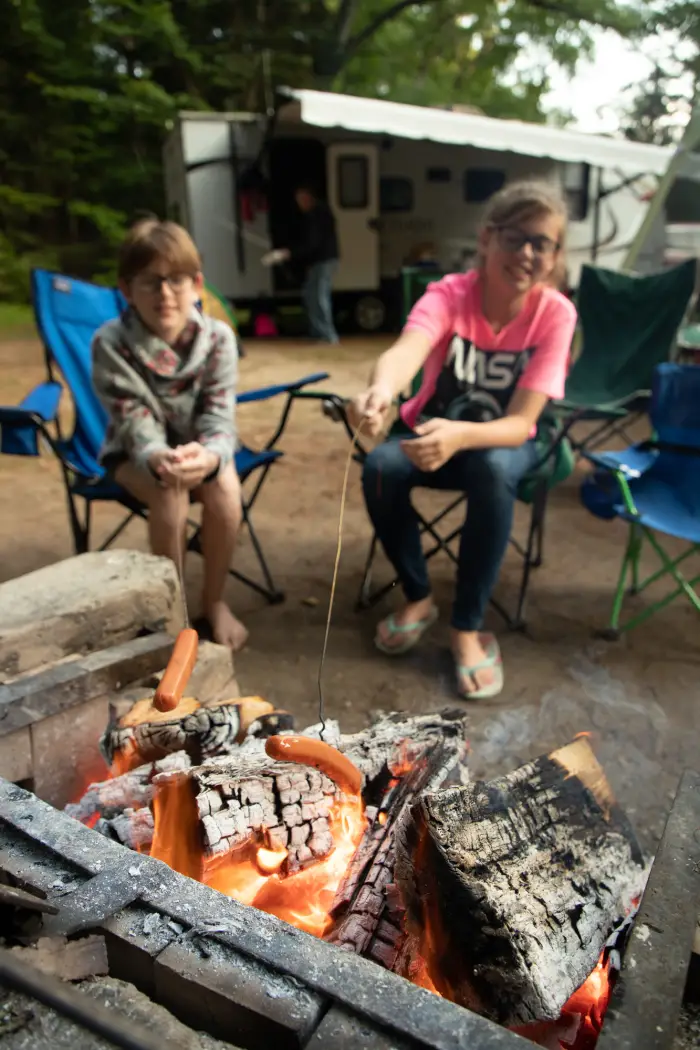 Two children sit by a campfire while using sticks to roast hotdogs